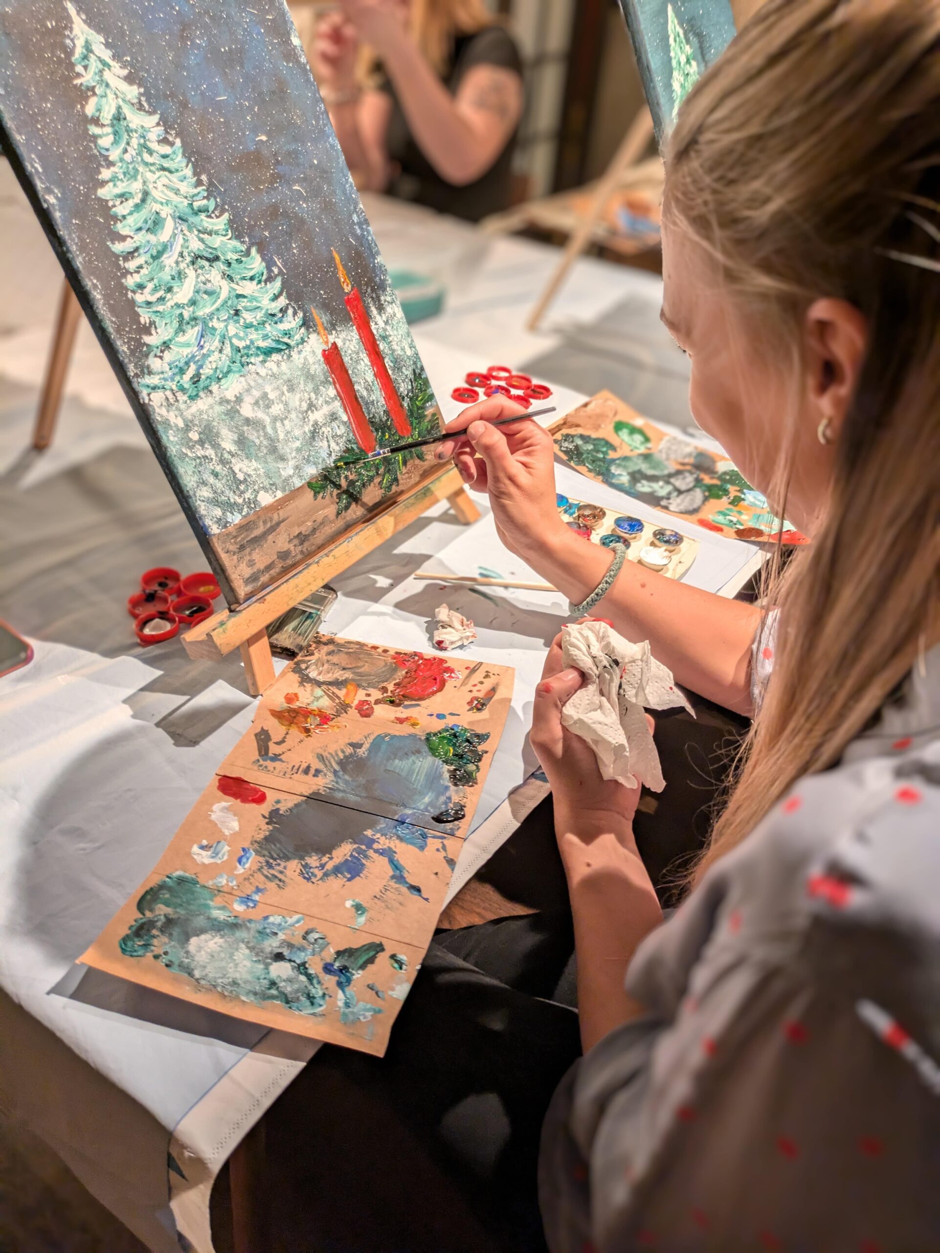 Kvinna målar vintermotiv med granar och röda ljus på duk vid målarworkshop